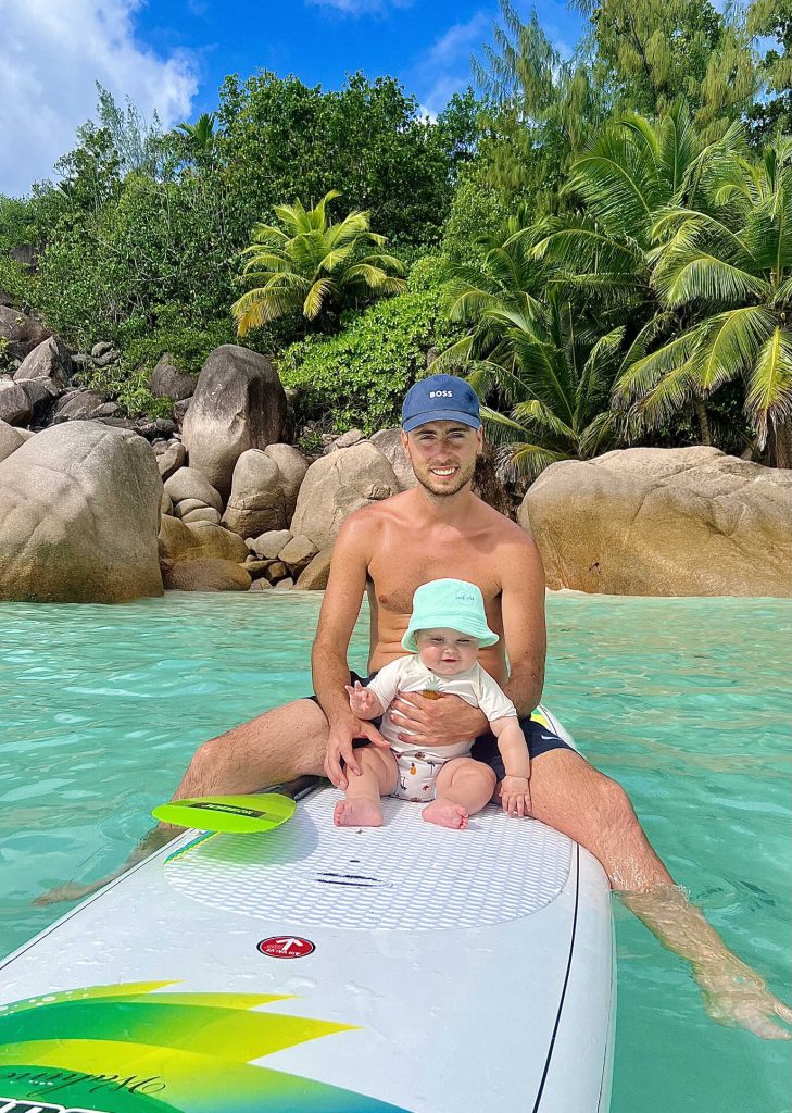 Père et enfant sur une planche dans une eau turquoise aux Seychelles, entourés de rochers et de végétation tropicale